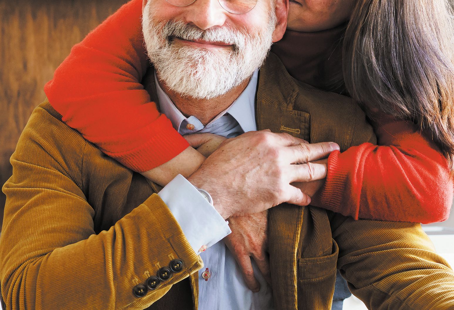 A woman in a red sweater hugs a man in a brown corduroy jacket, who smiles at the camera.
